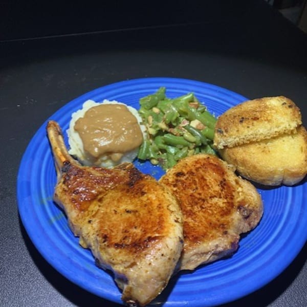 Two golden-brown pork chops, mashed potatoes with gravy, green beans, and toasted bread on a blue plate.