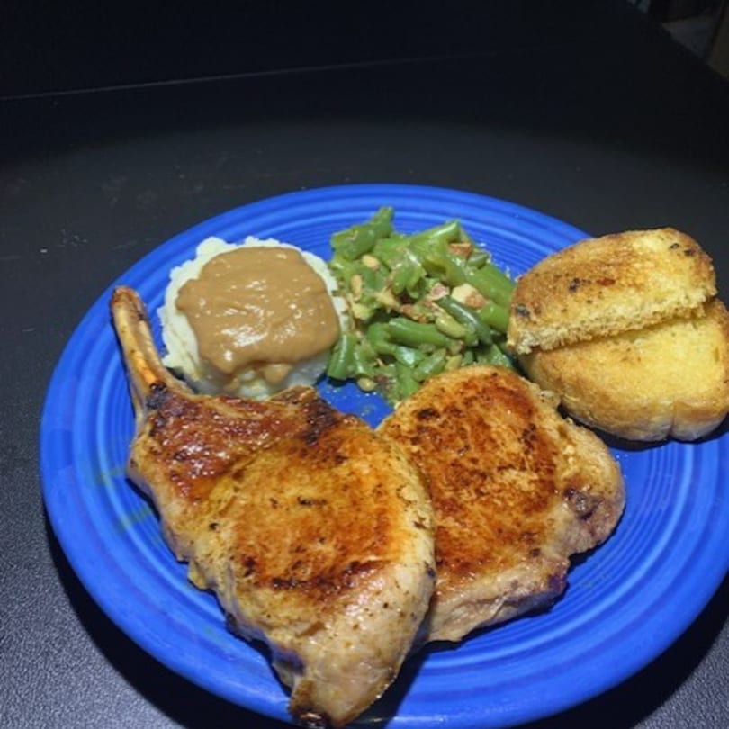 Two golden-brown pork chops, mashed potatoes with gravy, green beans, and toasted bread on a blue plate.