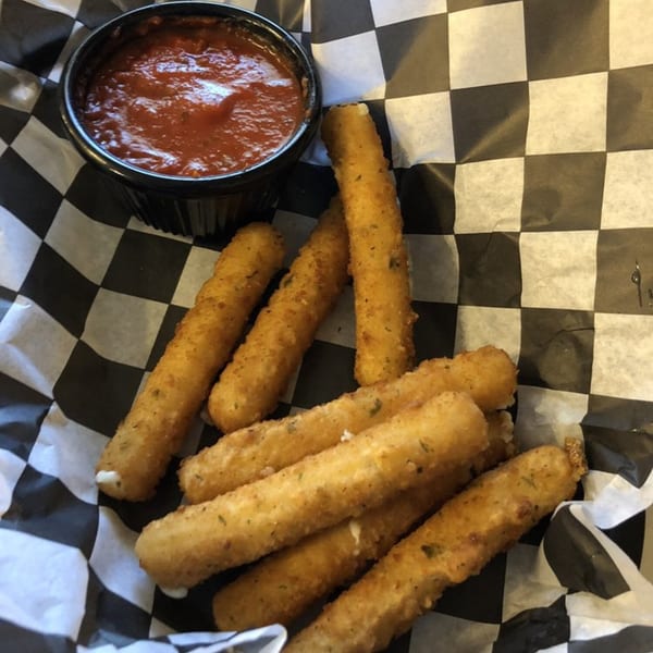 Golden-brown mozzarella sticks with marinara sauce in a black cup, served on checkered paper.