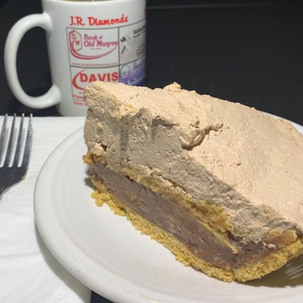 A slice of chocolate pie with a thick whipped topping on a white plate next to a JR Diamonds mug.